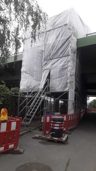 Hohes, mit Folie eingehaustes Gerüst unter einem Brückenbauwerk, davor Absperrbaken und ein Treppenaufgang.