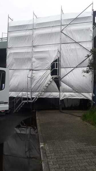 Mit Plane geschlossenes Gerüst an einer Fassade, davor ein Treppenaufgang und ein Transporter.