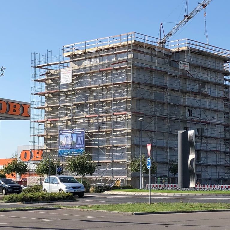Mehrgeschossiger Rohbau, vollständig eingerüstet, dahinter ein Baukran vor blauem Himmel.